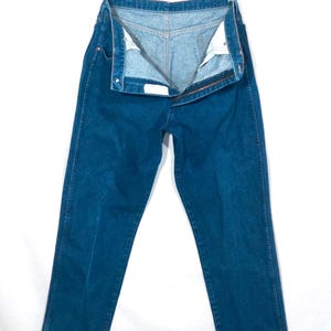 May include: A pair of open, dark blue denim jeans hanging on a metal hanger. The jeans are unbuttoned and unzipped, revealing the light blue interior. The jeans have a classic five-pocket design and a straight leg cut.