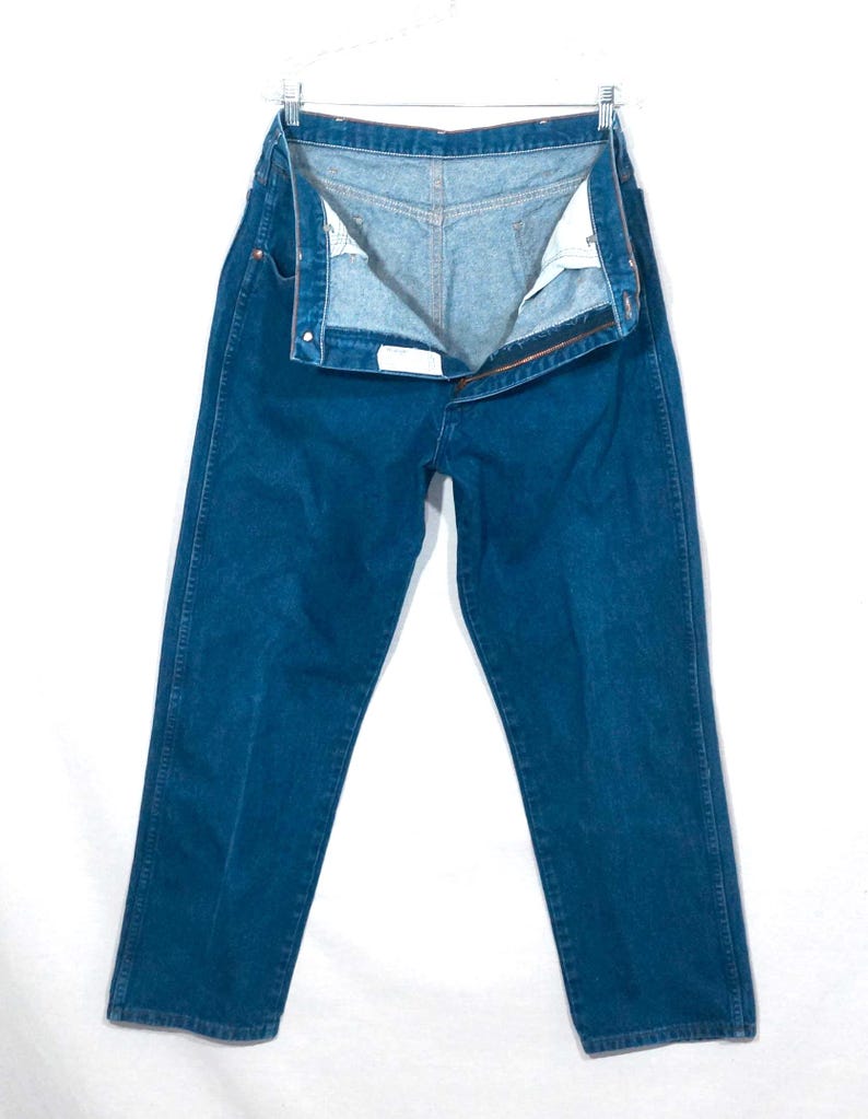 May include: A pair of open, dark blue denim jeans hanging on a metal hanger. The jeans are unbuttoned and unzipped, revealing the light blue interior. The jeans have a classic five-pocket design and a straight leg cut.