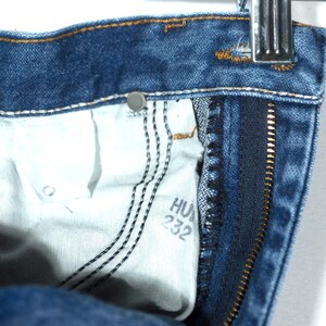 Puede incluir: Primer plano de unos vaqueros de mezclilla azules, que muestran el bolsillo interior y la cremallera. El forro del bolsillo es blanco, con costuras negras y el texto "HU 232". Los vaqueros est&aacute;n colgados de una percha met&aacute;lica.