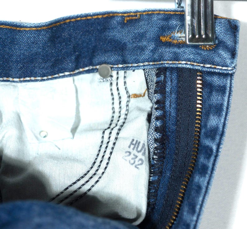 Puede incluir: Primer plano de unos vaqueros de mezclilla azules, que muestran el bolsillo interior y la cremallera. El forro del bolsillo es blanco, con costuras negras y el texto "HU 232". Los vaqueros est&aacute;n colgados de una percha met&aacute;lica.