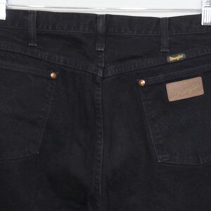 May include: Close-up of the back of black Wrangler jeans. The jeans have two back pockets, brass rivets, and a brown leather patch with the Wrangler logo. The stitching is visible, and the jeans are hanging from a metal hanger.