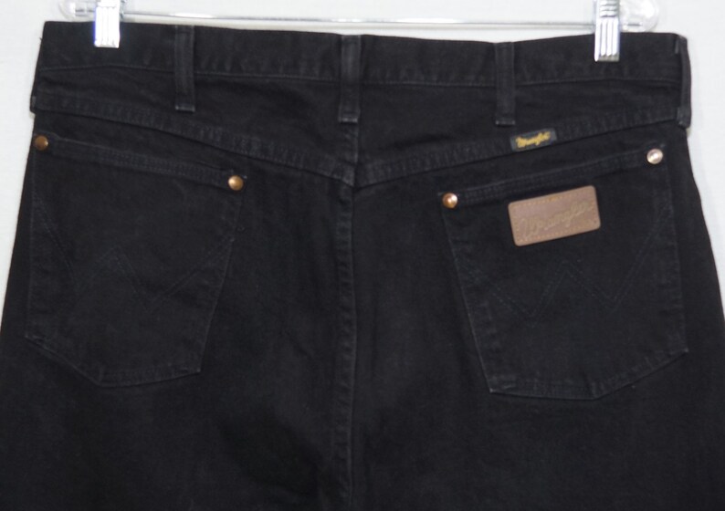 May include: Close-up of the back of black Wrangler jeans. The jeans have two back pockets, brass rivets, and a brown leather patch with the Wrangler logo. The stitching is visible, and the jeans are hanging from a metal hanger.