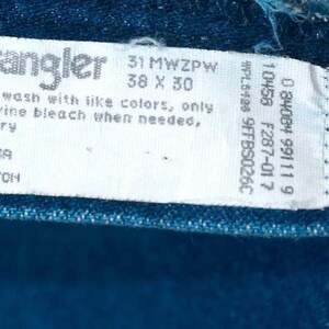 May include: Close-up of a blue denim tag from Wrangler jeans. The tag displays the brand name, size information (38 x 30 inches), and care instructions. The tag also states "MADE IN USA" and "100% COTTON".