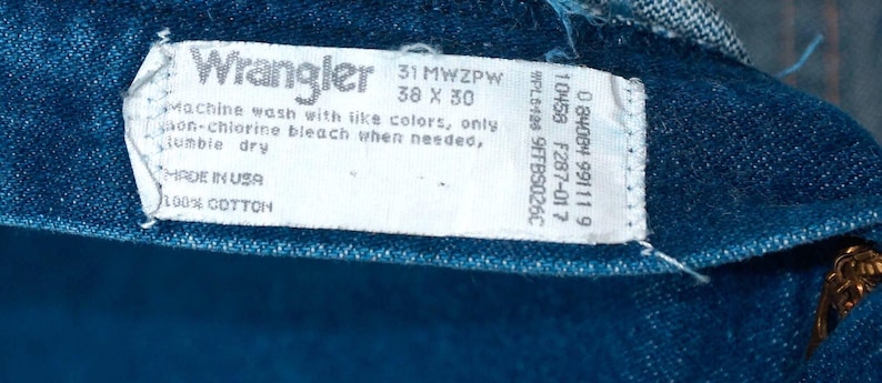 May include: Close-up of a blue denim tag from Wrangler jeans. The tag displays the brand name, size information (38 x 30 inches), and care instructions. The tag also states "MADE IN USA" and "100% COTTON".