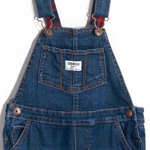 May include: Blue denim overalls with adjustable straps and metal hardware. The straps feature a red and black plaid pattern. The front pocket has the "OshKosh B'gosh" logo. The overalls have multiple pockets and are designed for children.