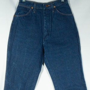 May include: Dark blue denim jeans with a high waist, featuring classic five-pocket styling. The jeans have copper-colored rivets and a button closure. The garment is hanging on a clear hanger against a white background.