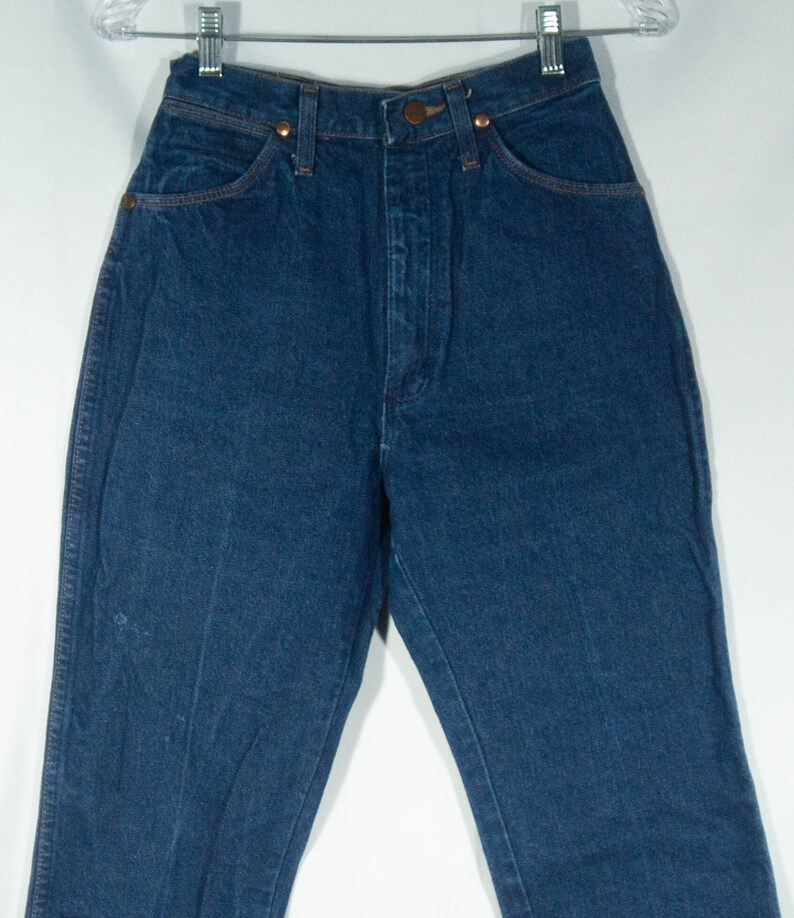 May include: Dark blue denim jeans with a high waist, featuring classic five-pocket styling. The jeans have copper-colored rivets and a button closure. The garment is hanging on a clear hanger against a white background.