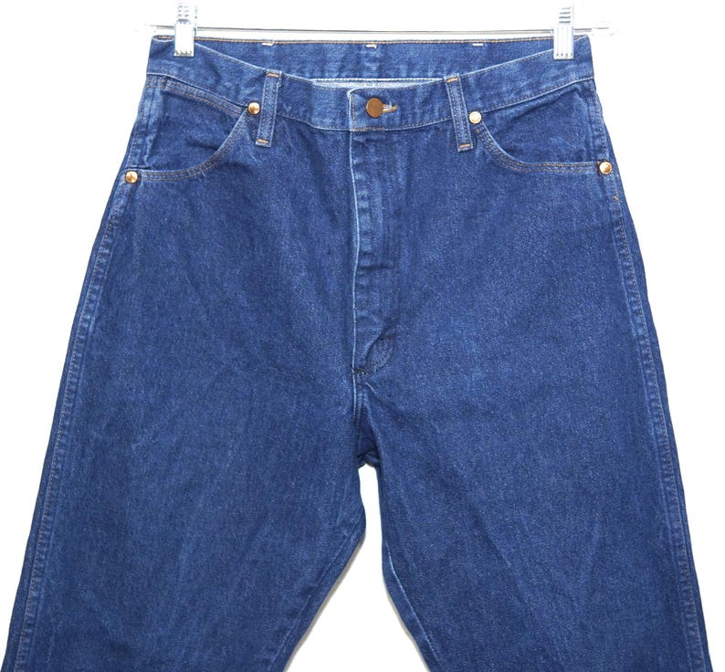 May include: A close-up shot of blue denim jeans. The jeans feature a classic five-pocket design, a button closure, and copper-coloured rivets. The jeans are suspended from a metal hanger against a white backdrop.