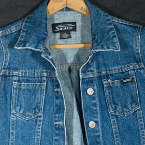 May include: A medium-wash denim jacket with a classic collar and button-up front. The jacket features two chest pockets and the brand name "Squeeze" on the label and pocket. The jacket is hanging on a wooden hanger.