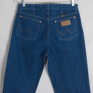 May include: Dark blue denim jeans with copper rivets and stitching. The back pockets feature a signature 'W' design. A brown leather patch with the brand name is visible on the back waistband. The jeans are hanging on a metal hanger.