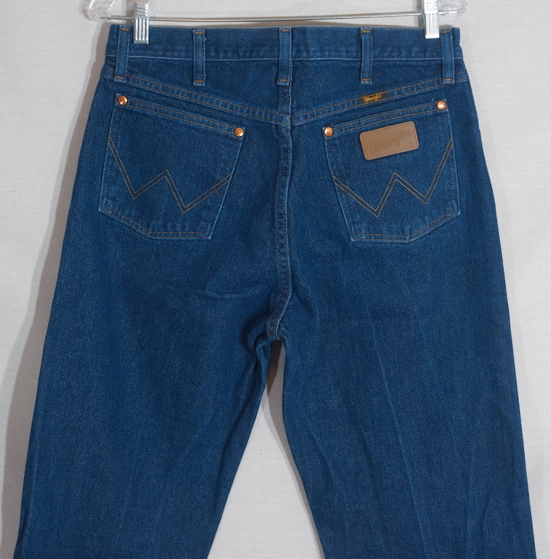May include: Dark blue denim jeans with copper rivets and stitching. The back pockets feature a signature 'W' design. A brown leather patch with the brand name is visible on the back waistband. The jeans are hanging on a metal hanger.