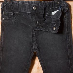 May include: Close-up of a pair of black denim pants. The pants have a button and zipper closure, with a small pocket on the front. The waistband is elasticated. The denim has a slightly faded appearance.