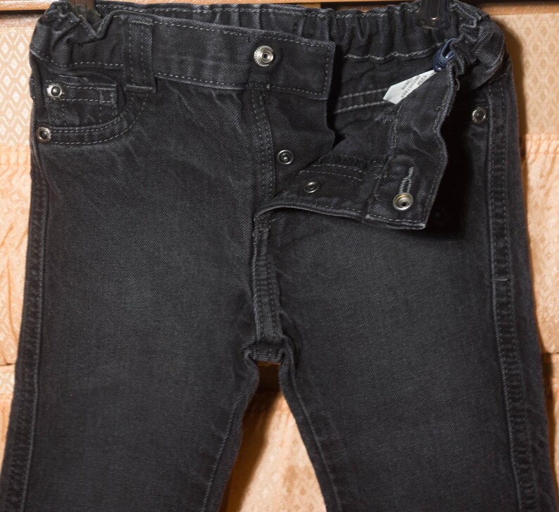 May include: Close-up of a pair of black denim pants. The pants have a button and zipper closure, with a small pocket on the front. The waistband is elasticated. The denim has a slightly faded appearance.