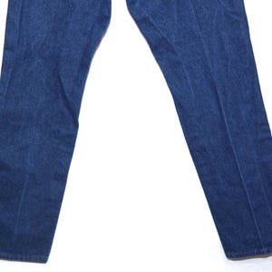 May include: Close-up of blue denim jeans. The jeans are a medium wash with a classic five-pocket design. The fabric appears sturdy and well-worn, with subtle fading and creases. The legs are straight cut.