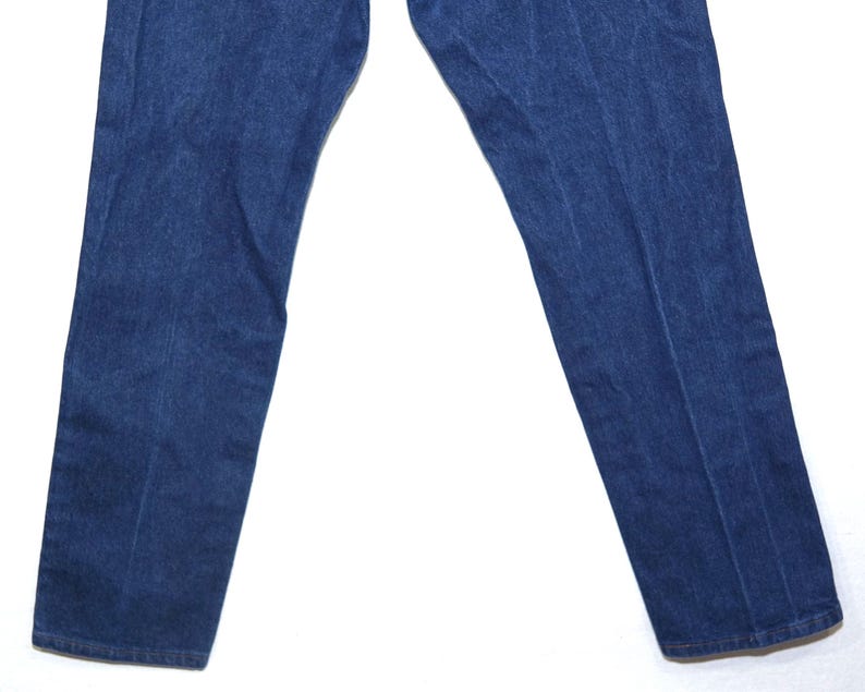 May include: Close-up of blue denim jeans. The jeans are a medium wash with a classic five-pocket design. The fabric appears sturdy and well-worn, with subtle fading and creases. The legs are straight cut.