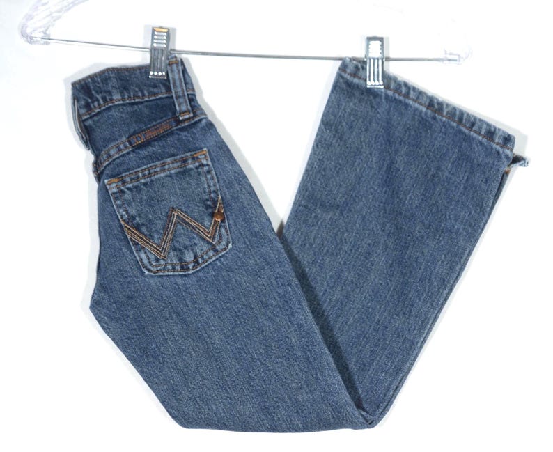 May include: Blue denim jeans with a classic five-pocket design, hanging from a metal hanger. The jeans feature a brown and gold stitched back pocket with a distinctive 'W' logo. The jeans are made of a durable denim material.