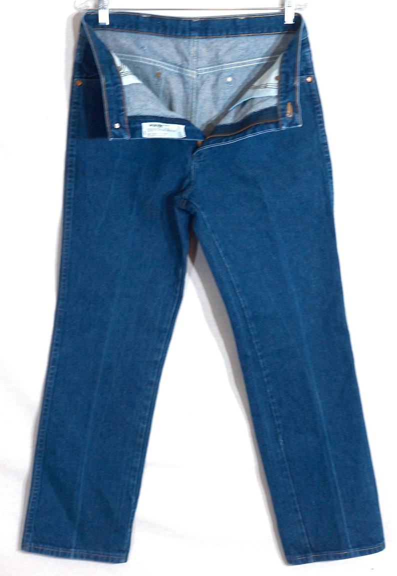 May include: A pair of blue denim jeans, unbuttoned and hanging from a metal hanger. The jeans have a classic five-pocket design and a straight leg cut. The denim is a medium wash, and the jeans appear to be unworn.