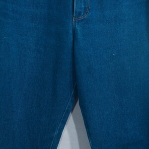 May include: Close-up of blue denim jeans. The jeans have a classic five-pocket design with a button and zipper closure. The denim is a medium wash, and the stitching is a contrasting gold color.