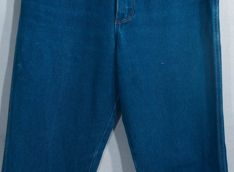 May include: Close-up of blue denim jeans. The jeans have a classic five-pocket design with a button and zipper closure. The denim is a medium wash, and the stitching is a contrasting gold color.