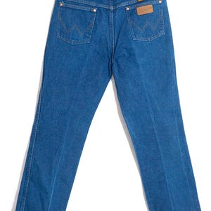 May include: Blue denim jeans with a classic five-pocket design. The jeans feature copper rivets, a brown leather patch, and the Wrangler logo. The back pockets have the signature W stitching. The jeans are hanging on a hanger.