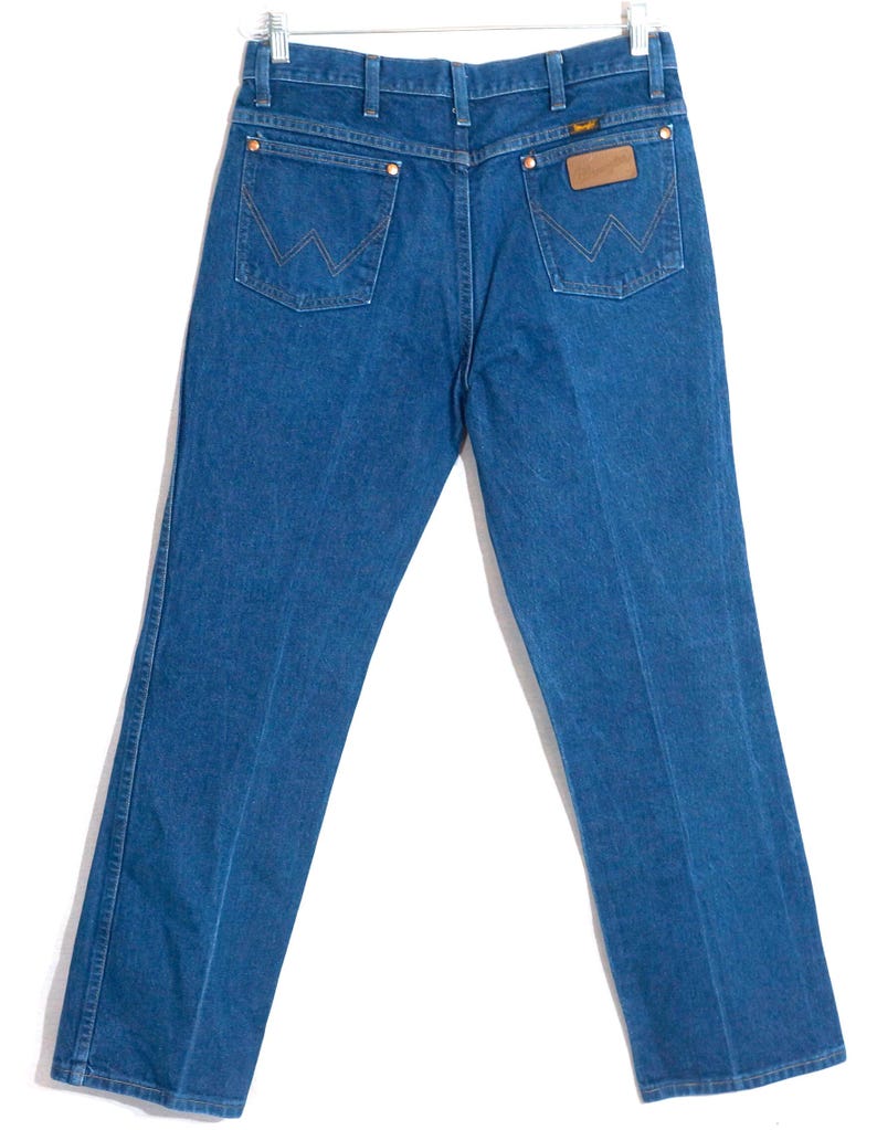 May include: Blue denim jeans with a classic five-pocket design. The jeans feature copper rivets, a brown leather patch, and the Wrangler logo. The back pockets have the signature W stitching. The jeans are hanging on a hanger.