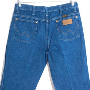 May include: Blue denim jeans with the Wrangler logo on the back pocket. The jeans have a classic five-pocket design with copper rivets and a straight leg. The back pockets feature the signature Wrangler "W" stitching.