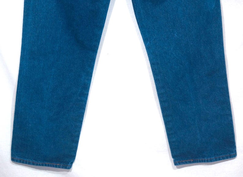 May include: Close-up of blue denim jeans. The jeans have a classic straight leg cut and a medium wash. The fabric appears to be sturdy, with visible texture. The hem is finished with a simple, clean stitch.