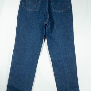 May include: Dark blue denim jeans with a classic five-pocket design. Features include copper rivets, a brown leather patch with the Wrangler logo, and signature stitching on the back pockets. The jeans are hung on a hanger.