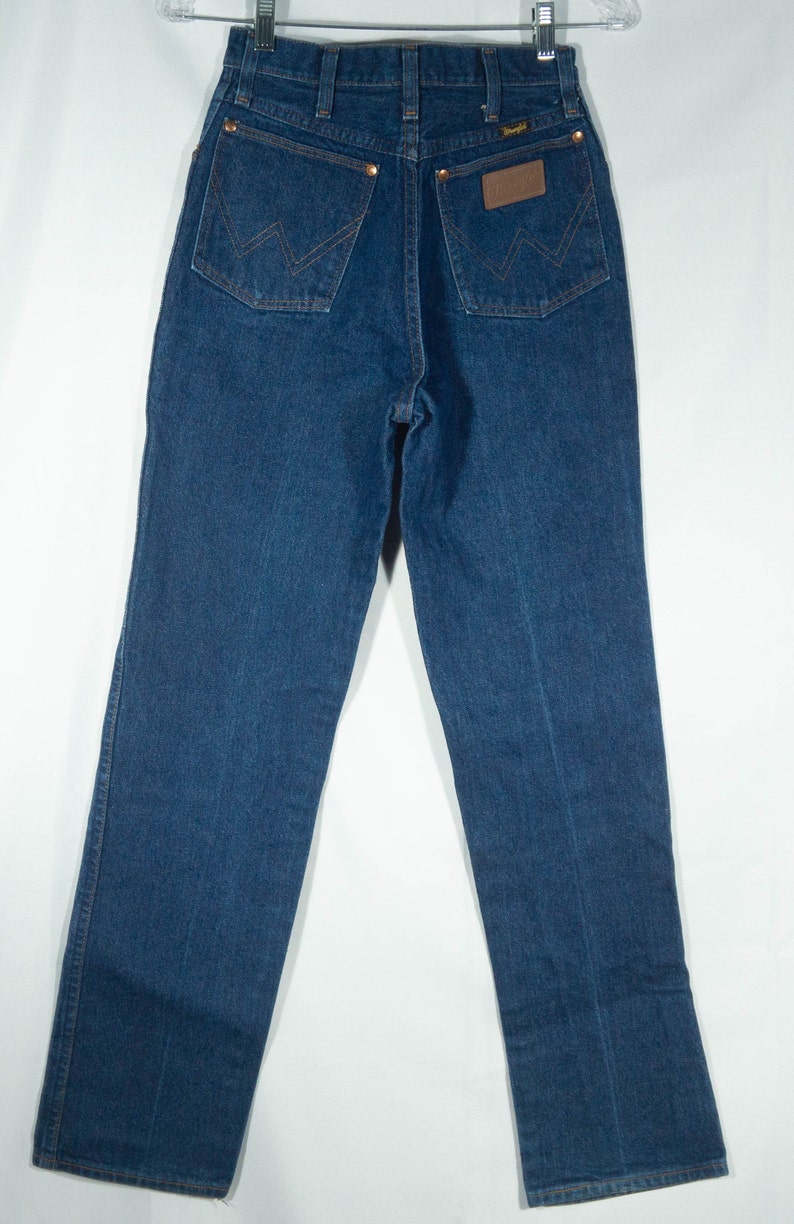 May include: Dark blue denim jeans with a classic five-pocket design. Features include copper rivets, a brown leather patch with the Wrangler logo, and signature stitching on the back pockets. The jeans are hung on a hanger.