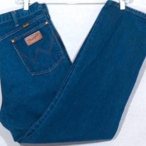 May include: Dark blue denim jeans with a classic five-pocket design. The jeans feature a brown leather patch on the back pocket with the brand name and a signature stitching detail. The jeans are hanging on a white hanger.