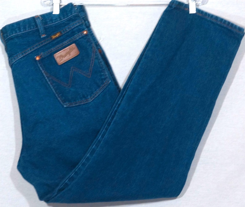 May include: Dark blue denim jeans with a classic five-pocket design. The jeans feature a brown leather patch on the back pocket with the brand name and a signature stitching detail. The jeans are hanging on a white hanger.