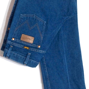 May include: Dark blue denim jeans with a classic five-pocket design, hanging on a wooden hanger. The jeans feature a brown leather patch and copper rivets. The back pocket has a distinctive stitching pattern.