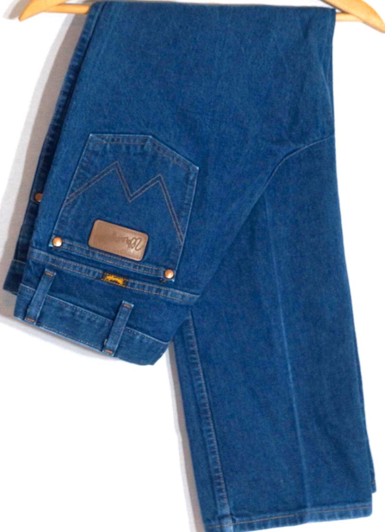 May include: Dark blue denim jeans with a classic five-pocket design, hanging on a wooden hanger. The jeans feature a brown leather patch and copper rivets. The back pocket has a distinctive stitching pattern.