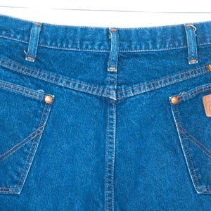 May include: Close-up of the back of blue denim jeans with orange stitching. The jeans have two back pockets with a signature "W" design and a brown leather "Wrangler" patch. The jeans are hanging from a metal hanger.