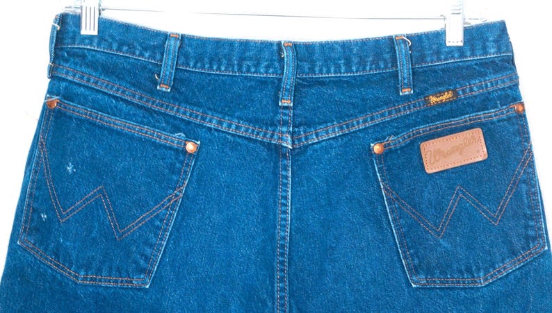 May include: Close-up of the back of blue denim jeans with orange stitching. The jeans have two back pockets with a signature "W" design and a brown leather "Wrangler" patch. The jeans are hanging from a metal hanger.