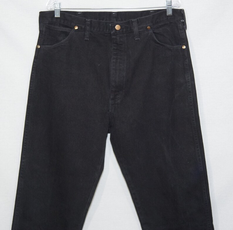 May include: Black denim jeans with a classic five-pocket design. The jeans have a button and zipper closure, and brass rivets. The jeans are hanging on a white background.