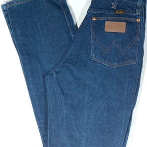 May include: Blue denim jeans with a classic five-pocket design. Features a brown leather patch and copper rivets. The jeans are hanging on a metal hanger. A vintage style.