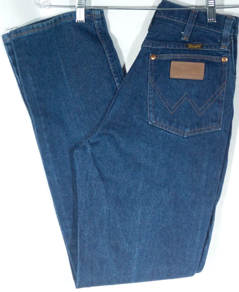 May include: Blue denim jeans with a classic five-pocket design. Features a brown leather patch and copper rivets. The jeans are hanging on a metal hanger. A vintage style.