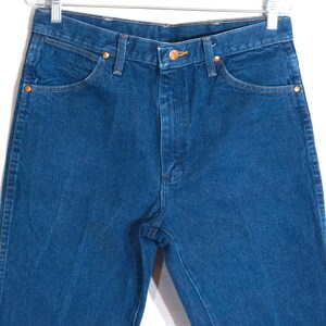 May include: Close-up of blue denim jeans with copper-coloured rivets and button. The jeans have a classic five-pocket design and a straight leg cut. The jeans are hanging on a metal hanger.