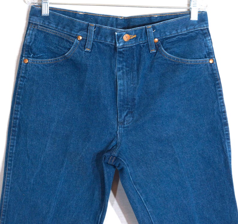 May include: Close-up of blue denim jeans with copper-coloured rivets and button. The jeans have a classic five-pocket design and a straight leg cut. The jeans are hanging on a metal hanger.