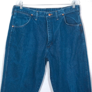 May include: Close-up of a pair of blue denim jeans. The jeans have a classic five-pocket design, copper rivets, and a button closure. The denim is a medium wash, and the jeans are hanging on a clear hanger.