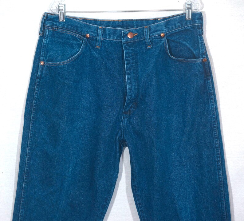May include: Close-up of a pair of blue denim jeans. The jeans have a classic five-pocket design, copper rivets, and a button closure. The denim is a medium wash, and the jeans are hanging on a clear hanger.