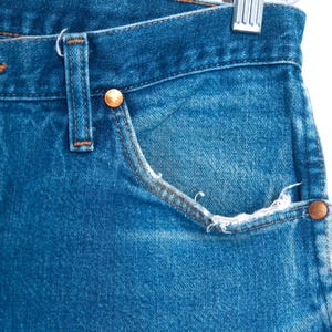 Puede incluir: Primer plano de unos vaqueros de mezclilla azules con remaches de color cobre y un bolsillo. Los vaqueros muestran signos de desgaste, con bordes deshilachados en el bolsillo. La mezclilla es de lavado medio y los vaqueros cuelgan de una percha met&aacute;lica.