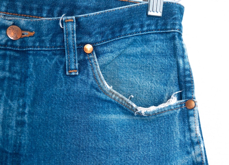 Puede incluir: Primer plano de unos vaqueros de mezclilla azules con remaches de color cobre y un bolsillo. Los vaqueros muestran signos de desgaste, con bordes deshilachados en el bolsillo. La mezclilla es de lavado medio y los vaqueros cuelgan de una percha met&aacute;lica.