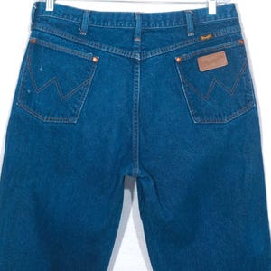 May include: Dark blue denim jeans with the Wrangler logo on the back pocket. The jeans have classic five-pocket styling, copper rivets, and a straight leg. The jeans are hanging on a metal hanger.
