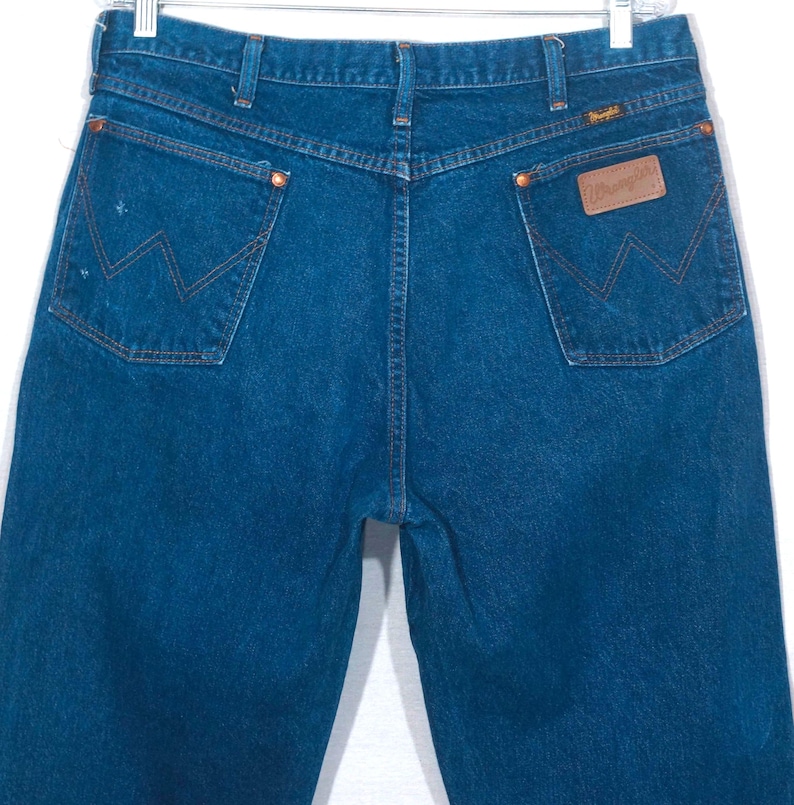 May include: Dark blue denim jeans with the Wrangler logo on the back pocket. The jeans have classic five-pocket styling, copper rivets, and a straight leg. The jeans are hanging on a metal hanger.