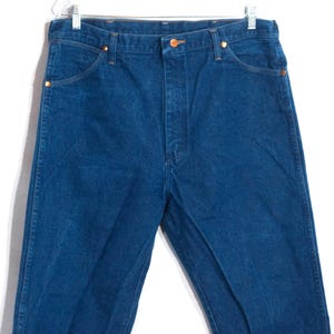 May include: A pair of blue denim jeans with a classic five-pocket design. The jeans have a button and zipper closure, and copper-colored rivets. The jeans are hanging on a metal hanger against a white background.