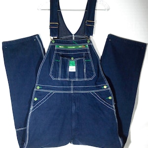 Könnte beinhalten: Dunkelblaue Jeans-Latzhose mit weißen Nähten und grünen Akzenten. Merkmale sind verstellbare Träger, mehrere Taschen und grüne Knopfverschlüsse. Die Latzhose hängt an einem transparenten Kleiderbügel.