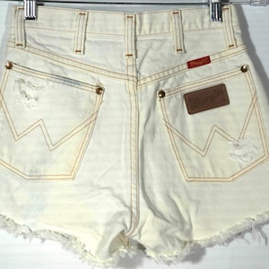 May include: Cream-colored denim shorts with a frayed hem, featuring the Wrangler logo on the back pocket. The shorts have a high waist and distressed details on the pockets. The stitching is a contrasting tan color.