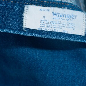 May include: Close-up of blue denim jeans with a white label. The label reads "Wrangler" and includes washing instructions and size 12. Made in the USA.